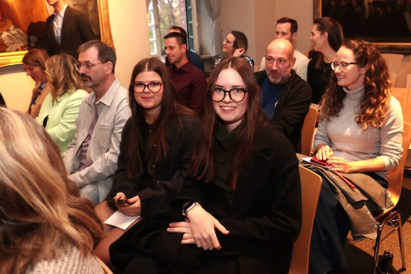 Eine Gruppe von Menschen sitzt bei einer Veranstaltung in einer Halle in Reihen. Die meisten sind leger gekleidet. Zwei junge Frauen mit langen braunen Haaren und Brillen sitzen in der Mitte im Vordergrund und lächeln in die Kamera. Andere blicken nach vorne, beschäftigt und aufmerksam. An den Wänden hängen Gemälde.
