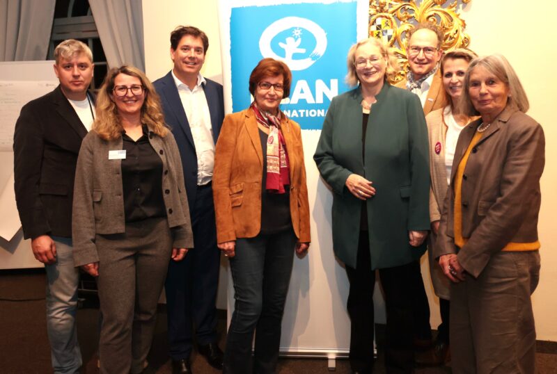 Acht Erwachsene, fünf Frauen und drei Männer, stehen lächelnd in einem Halbkreis in einem Raum. Hinter ihnen steht eine Tafel mit einem blauen Schild mit der Aufschrift "ICAN International". Sie sind geschäftlich oder leger gekleidet und scheinen an einer Veranstaltung oder einem Treffen teilzunehmen.