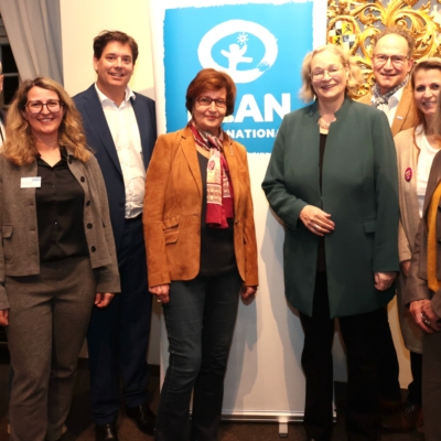 Acht Erwachsene stehen in einer Reihe und lächeln in die Kamera vor einem blau-weißen ICAN International-Schild. Sie tragen Geschäfts- oder Freizeitkleidung, einige von ihnen Jacken und Schals. Der Schauplatz scheint ein Innenraum zu sein, in dem verziertes und modernes Dekor zu sehen ist.