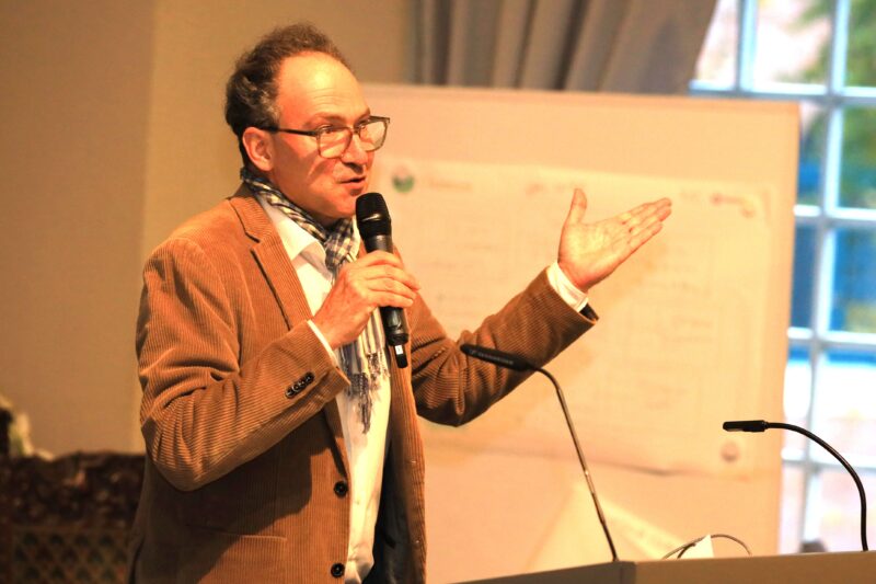 Ein Mann mittleren Alters mit Brille und braunem Blazer spricht auf einem Podium in ein Mikrofon und gestikuliert mit seiner linken Hand. Er trägt einen gemusterten Schal und steht vor einem verschwommenen Whiteboard und einem hohen Fenster, durch das Licht fällt.