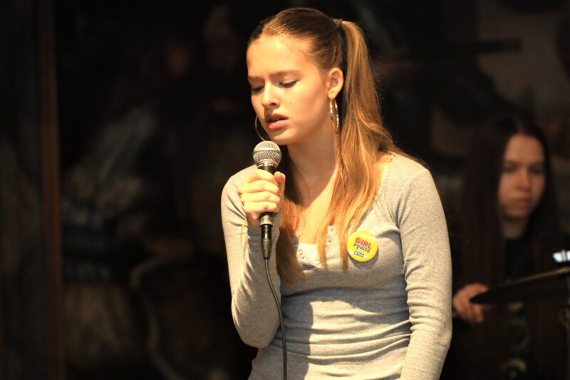 Eine junge Frau mit langen, hellbraunen Haaren singt mit geschlossenen Augen in ein Mikrofon. Sie trägt ein graues Hemd und eine runde Girl Power-Anstecknadel. Im Hintergrund ist eine verschwommene Figur zu sehen, die Schlagzeug spielt, vor einem dunklen, undeutlichen Hintergrund.