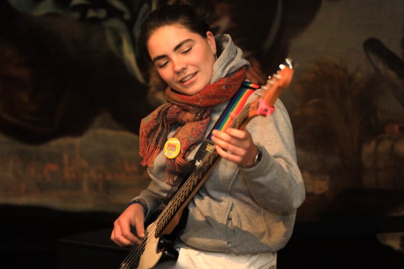 Eine junge Person mit zurückgebundenen dunklen Haaren, die einen grauen Kapuzenpulli, einen rot gemusterten Schal und eine Anstecknadel mit der Aufschrift "Queer Power" trägt, spielt eine elektrische Bassgitarre. Ihre Augen sind geschlossen und sie lächeln, konzentriert auf die Musik. Der Hintergrund ist schummrig und unscharf.