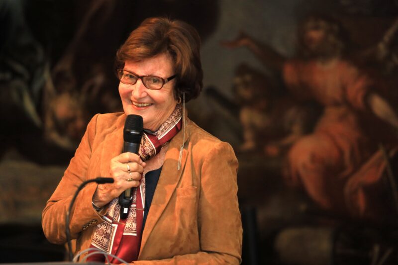 Eine ältere Frau mit kurzen braunen Haaren und einer Brille lächelt freundlich und hält ein Mikrofon in der Hand. Sie trägt eine braune Jacke, einen gemusterten Schal und einen goldenen Ring. Der Hintergrund ist schummrig und zeigt ein klassisches Kunstwerk mit weichen, gedämpften Farben.