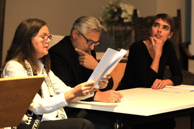 Drei Personen sitzen an einem Tisch und diskutieren. Die Frau auf der linken Seite liest aus Papieren, der Mann in der Mitte und die Frau auf der rechten Seite stützen beide eine Hand auf ihr Kinn und hören aufmerksam zu. Auf dem Tisch liegen weitere Papiere und im Hintergrund steht ein Blumengesteck.