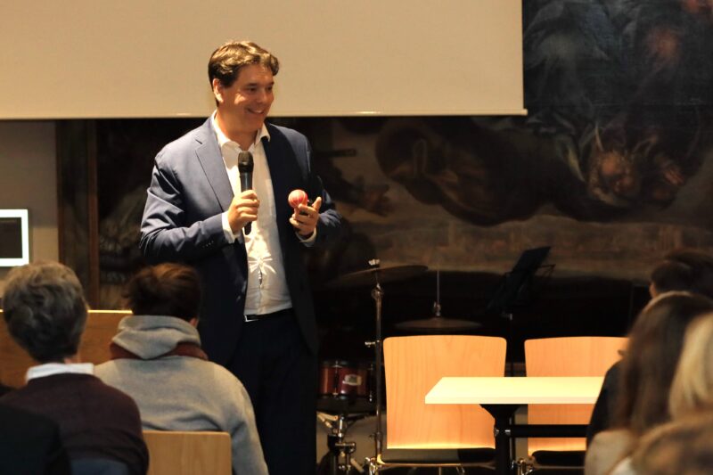 Ein Mann in einem blauen Anzug hält ein Mikrofon und einen roten Apfel in der Hand, während er lächelnd zu einem auf Stühlen sitzenden Publikum spricht. Hinter ihm stehen ein Schlagzeug und ein Notenständer. Der Raum ist warm beleuchtet und an der Wand befindet sich ein großes Wandgemälde.