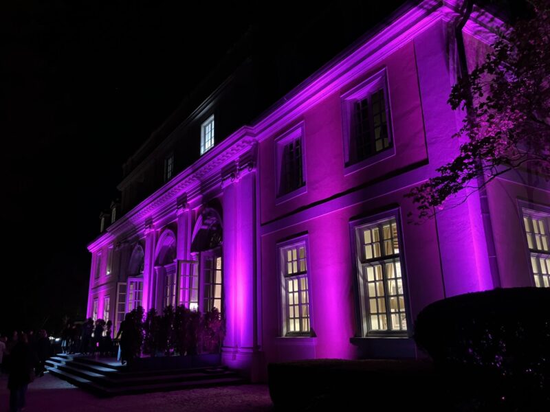 Ein großes, elegantes Gebäude wird nachts mit leuchtenden violetten Lichtern angestrahlt. Die helle Beleuchtung hebt die Fenster, Säulen und architektonischen Merkmale des Gebäudes hervor. In der Nähe des Eingangs auf der linken Seite sind Schatten und Silhouetten von Menschen zu sehen.