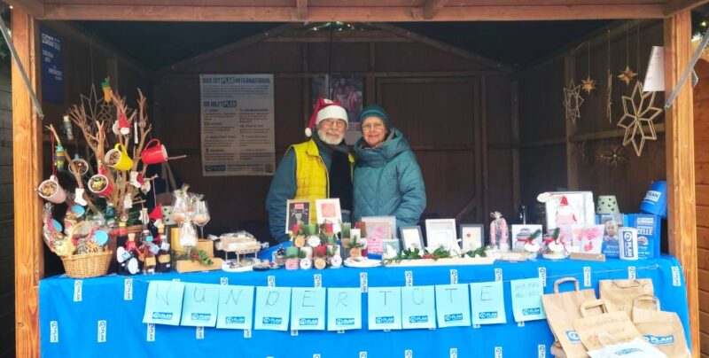 Zwei lächelnde Menschen stehen hinter einem festlichen Marktstand mit einer blauen Tischdecke mit der Aufschrift "Wundertüte". Auf dem Tisch stehen Kunsthandwerk, gerahmte Kunstwerke, Leckereien und Gläser. Der hölzerne Stand ist mit Sternen, hängenden Bechern und Weihnachtsschmuck dekoriert. Eine Person trägt eine Weihnachtsmannmütze.