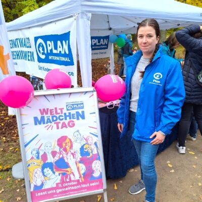 Eine Frau in einer blauen Jacke von Plan International steht neben einem Plakat für den "Welt-Mädchentag" mit rosa Luftballons und illustrierten Mädchen. Hinter ihr steht ein weißes Zelt mit Plan International-Bannern und darunter versammelten Menschen.