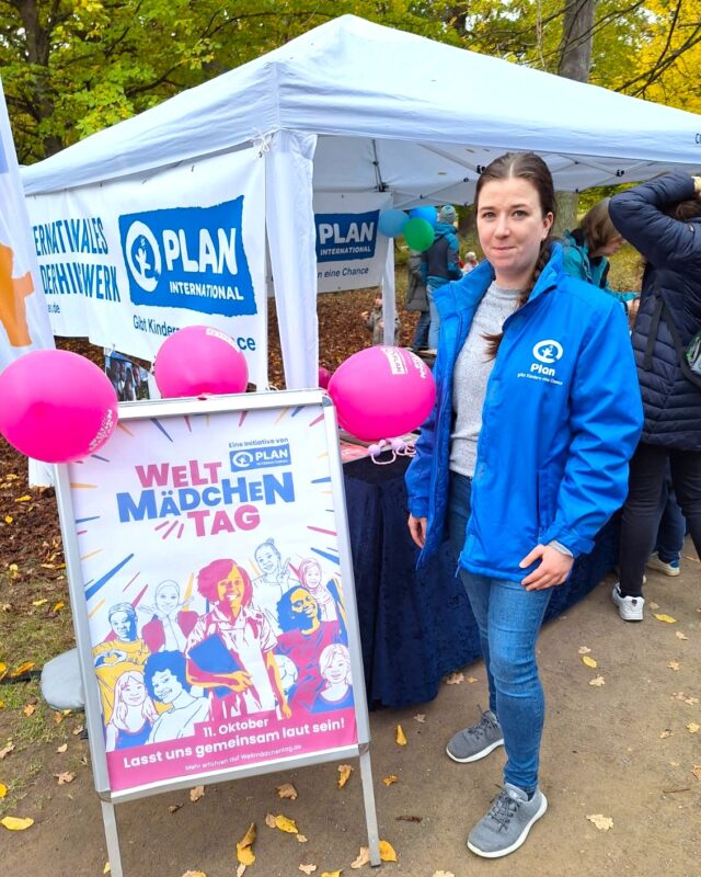 Eine Frau in einer blauen Jacke von Plan International steht neben einem Plakat für den "Welt-Mädchentag" mit rosa Luftballons und illustrierten Mädchen. Hinter ihr steht ein weißes Zelt mit Plan International-Bannern und darunter versammelten Menschen.