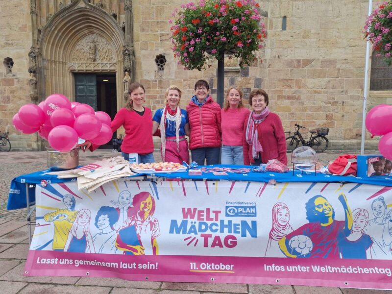 Fünf Frauen in rosa und roter Kleidung stehen hinter einem Tisch im Freien, der mit rosa Luftballons, Broschüren, Tragetaschen und Leckereien dekoriert ist. Ein blaues Banner mit der Aufschrift "Internationaler Tag des Mädchens" ist zu sehen, im Hintergrund ein historisches Steingebäude.