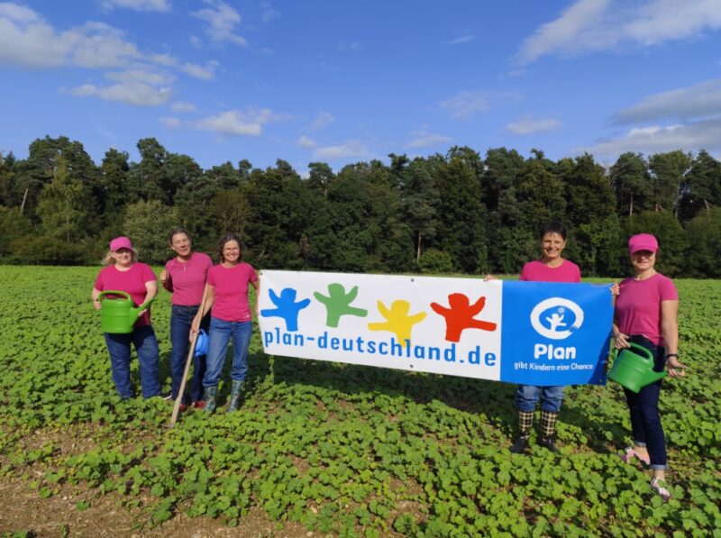 Fünf Frauen in rosa Hemden und Hüten stehen auf einer grünen Wiese und halten Gießkannen und ein großes Banner mit der Aufschrift plan-deutschland.de mit bunten menschlichen Figuren und dem blauen Plan International-Logo. Im Hintergrund sind Bäume und blauer Himmel zu sehen.