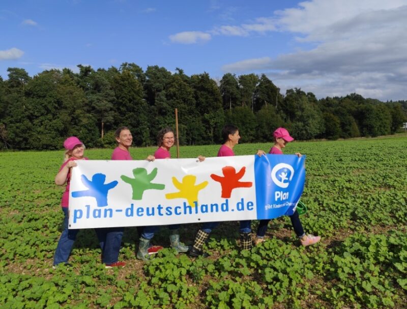 Vier Frauen in rosa Hemden laufen durch eine grüne Wiese und halten ein großes "plan-deutschland.de"-Banner mit bunten menschlichen Silhouetten und einem blauen Plan International-Logo. Im Hintergrund sind Wald und blauer Himmel mit Wolken zu sehen.