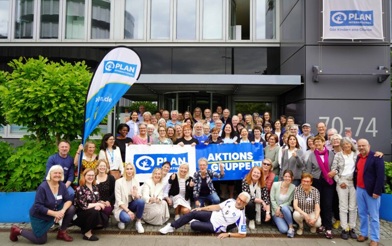 Eine große Gruppe verschiedener Erwachsener versammelt sich vor einem modernen Gebäude. Viele lächeln und halten blaue Transparente mit der Aufschrift PLAN und AKTIONS GRUPPEN. Links steht eine blaue Fahne mit dem Plan-Logo, und ein Schild am Gebäude zeigt PLAN International.