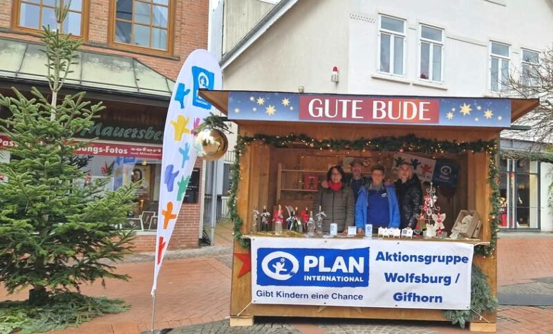 Eine hölzerne Weihnachtsbude mit der Aufschrift GUTE BUDE steht in einer Backsteinstraße. Vier Personen sitzen darin hinter Tischen mit Kunsthandwerk und Dekoration. Auf einem Transparent steht Plan International Aktionsgruppe Wolfsburg/Gifhorn. Auf der linken Seite stehen ein geschmückter Weihnachtsbaum und eine Fahne.
