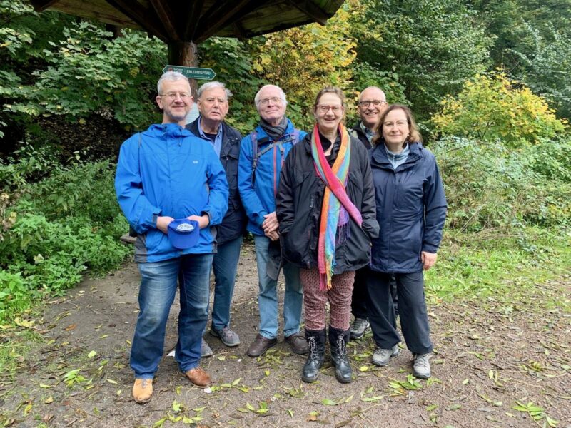 Sechs Erwachsene stehen dicht nebeneinander auf einem unbefestigten Weg, umgeben von grünem Laub. Die meisten tragen Jacken, einige mit Kapuzen, und eine Person im Vordergrund trägt einen bunten Schal. Sie alle lächeln in die Kamera unter einem hölzernen Schutzdach. Der Boden ist mit Blättern bedeckt.