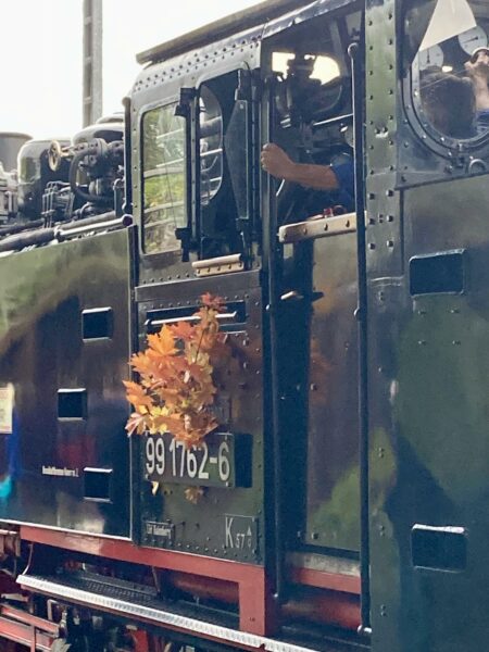 Nahaufnahme des Führerhauses einer schwarzen Dampflokomotive, an der neben der Nummer "99 1762-6" ein Strauß Herbstblätter befestigt ist. Im Inneren ist eine Person in einem blauen Hemd zu sehen, die den Zug bedient. Das verwitterte Metall des Zuges glänzt im natürlichen Tageslicht.