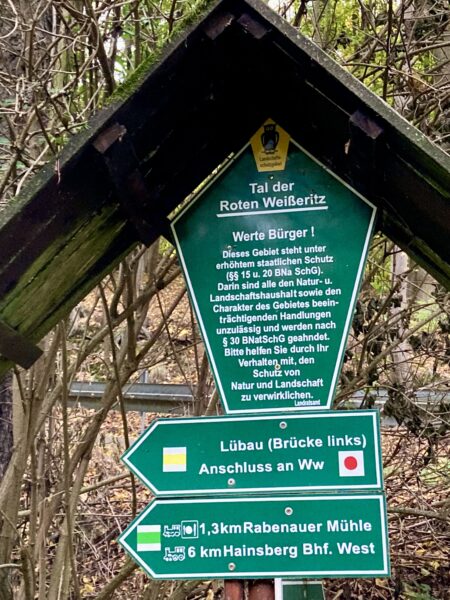 Eine Holzkonstruktion trägt drei grüne Wegweiser in deutscher Sprache. Das oberste Schild, das die Form eines Fünfecks hat, informiert über den Naturschutz im Tal der Roten Weißeritz. Darunter weisen zwei Pfeilschilder auf Orte hin: Rabenauer Mühle (1,3 km) und Hainsberg Bhf. West (6 km).