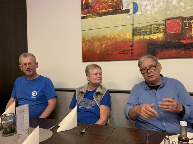 Drei ältere Erwachsene sitzen an einem mit gefalteten Servietten und Speisekarten gedeckten Restauranttisch. Der Mann auf der linken Seite und die Frau in der Mitte tragen blaue Hemden; der Mann auf der rechten Seite trägt eine Brille und gestikuliert beim Sprechen. Hinter ihnen hängt abstrakte Kunst an der Wand.