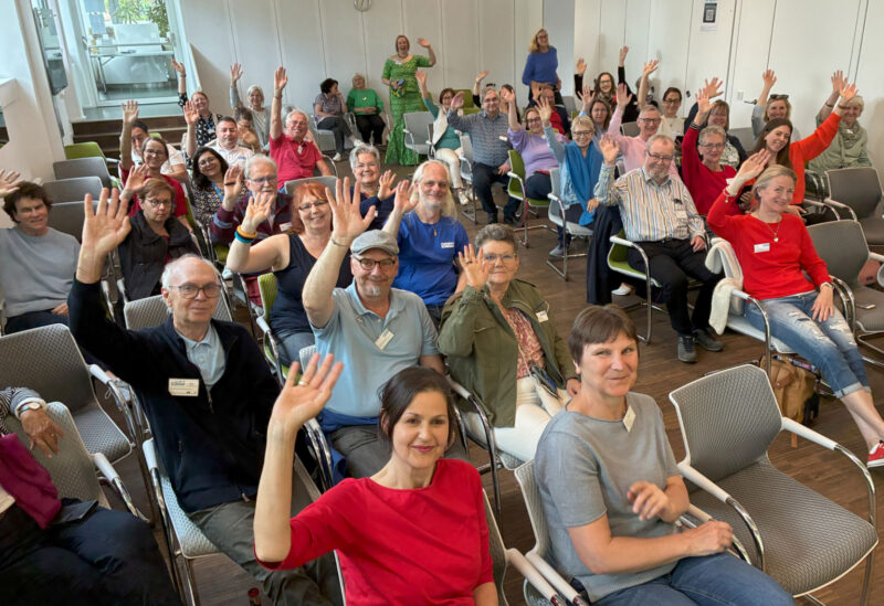 Eine Gruppe von etwa vierzig Erwachsenen unterschiedlichen Alters und Aussehens sitzt in Stuhlreihen in einem hellen Raum, lächelt und hebt eine Hand zum Gruß oder winkt in die Kamera. Einige tragen Namensschilder. Eine Frau steht im Hintergrund und winkt ebenfalls.