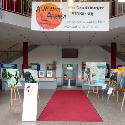 In der Lobby mit rotem Teppich sind auf Staffeleien Gemälde mit Tiermotiven ausgestellt, vor allem von Elefanten. Auf großen Bannern und einem großen Schild über dem Eingang wird für "Auf nach Afrika" geworben. Im Hintergrund sind Treppen und Hinweisschilder zu sehen.