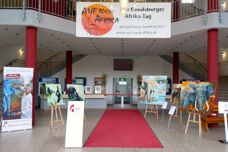 In der Lobby mit rotem Teppich sind auf Staffeleien Gemälde mit Tiermotiven ausgestellt, vor allem von Elefanten. Auf großen Bannern und einem großen Schild über dem Eingang wird für "Auf nach Afrika" geworben. Im Hintergrund sind Treppen und Hinweisschilder zu sehen.