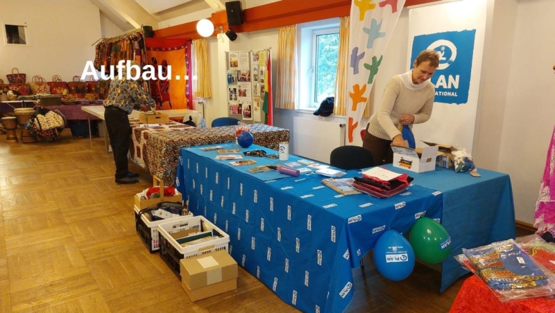 Zwei Personen bauen bei einer Veranstaltung in einer Halle Tische auf. Eine Frau arrangiert Materialien an einem Stand mit blauen Tischen, Oxfam-Postern und Luftballons. Eine andere Person steht an einem bunten Stand mit Taschen und Trommeln. Auf dem Bild erscheint das deutsche Wort "Aufbau...".