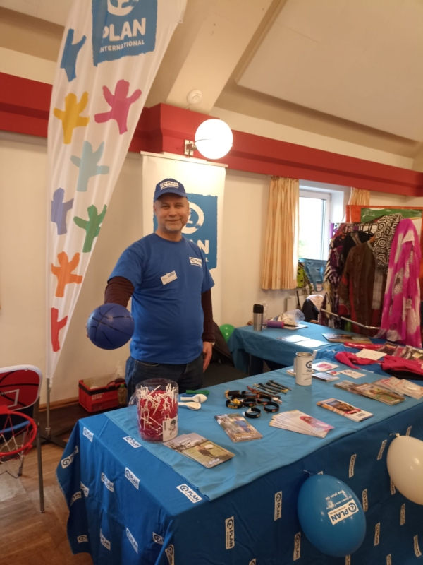 Ein lächelnder Mann in einem blauen Plan International-T-Shirt und einer Mütze steht hinter einem Tisch mit Broschüren, Armbändern, einer Spendendose und Luftballons. Er hält einen blauen Stressball in der Hand. Um den Stand herum sind bunte Gegenstände und eine große Plan International-Flagge angebracht.