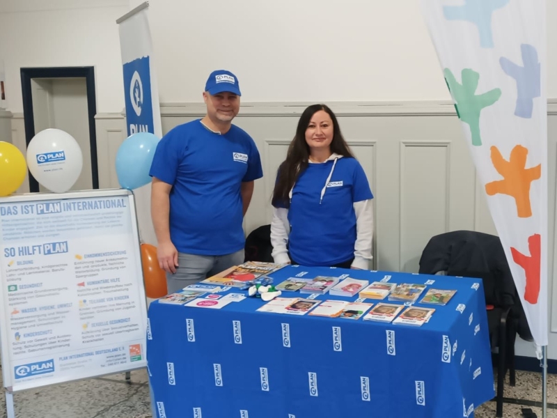 Zwei lächelnde Menschen in blauen Hemden stehen hinter einem Tisch, der mit Broschüren, Büchern und kleinen Gegenständen bedeckt ist. Auf dem Tisch und dem nahe gelegenen Banner ist das Logo von "Plan International" zu sehen. Bunte Luftballons und eine große Flagge mit abstrakten Figuren schmücken die Szene in einem hellen Raum.