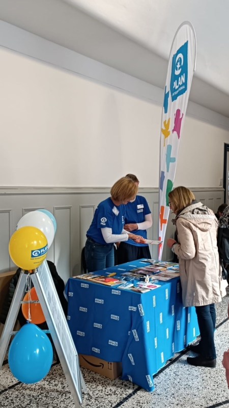 Zwei Frauen in blauen Hemden stehen hinter einem mit Broschüren bedeckten Werbetisch und unterhalten sich mit einer Frau in einem beigen Mantel. Auf dem Tisch liegen ein blaues Tuch mit einem Logo, bunte Luftballons, eine Schautafel und ein großes Transparent mit bunten Handabdrücken.