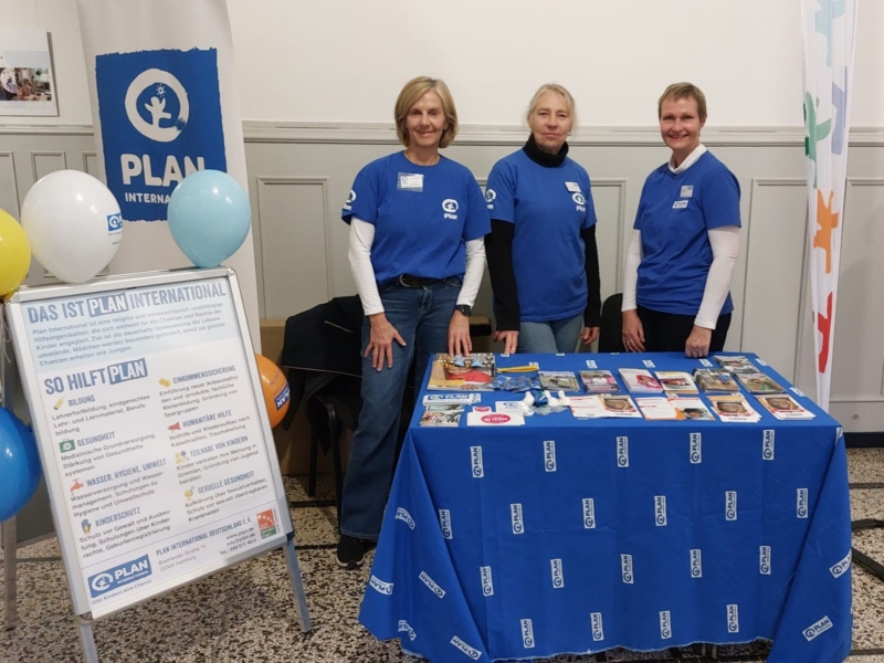 Drei Frauen stehen hinter einem Schautisch, der mit Broschüren und Flyern von Plan International bedeckt ist. Sie tragen blaue Hemden von Plan International und lächeln. Luftballons und ein Transparent zeigen das Logo. Ein Schild beschreibt den Auftrag und die Aktivitäten der Organisation auf Deutsch.