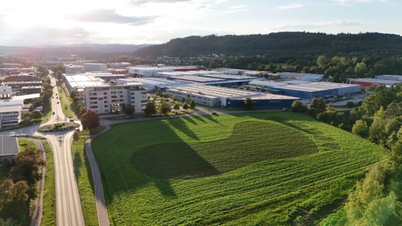 Luftaufnahme eines grünen Feldes mit einem großen herzförmigen Umriss in dunkleren Pflanzen. Um das Feld herum befinden sich Straßen, moderne Gebäude und Industrielagerhäuser. Bäume und ein hügeliges Waldgebiet erscheinen im Hintergrund unter einem teilweise bewölkten Himmel.