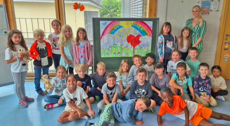 Eine Gruppe unterschiedlicher Kinder und eine Frau stehen und sitzen zusammen in einem Klassenzimmer und lächeln in die Kamera. Hinter ihnen ist eine bunte Zeichnung auf einer Tafel zu sehen, die einen Regenbogen, Herzen, Häuser und die Worte "Unsere Klasse" zeigt. Einige Kinder halten Stofftiere in der Hand.