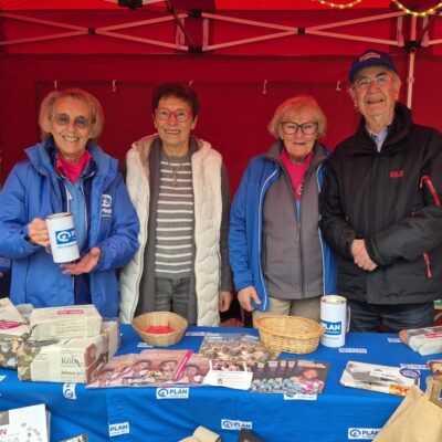Vier lächelnde ältere Erwachsene stehen hinter einem Tisch an einem Stand, der mit Broschüren, Spendendosen und kleinen Gegenständen von Plan International bedeckt ist. Zwei Frauen tragen blaue Jacken mit dem Plan-Logo, alle sind warm gekleidet. Im Hintergrund ist ein rotes Zeltdach zu sehen.
