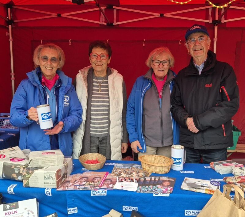 Vier lächelnde ältere Erwachsene stehen hinter einem Tisch an einem Stand, der mit Broschüren, Spendendosen und kleinen Gegenständen von Plan International bedeckt ist. Zwei Frauen tragen blaue Jacken mit dem Plan-Logo, alle sind warm gekleidet. Im Hintergrund ist ein rotes Zeltdach zu sehen.