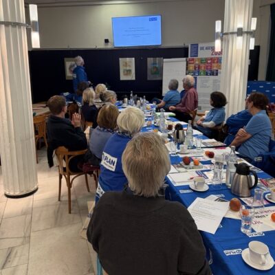 Eine Gruppe von Menschen sitzt um einen langen Tisch mit blauen Tischtüchern, Papieren, Obst und Kaffeekannen. Eine Person steht vorne und zeigt eine Diashow auf einer Leinwand. Der Raum ist mit Säulen ausgestattet, und die Teilnehmer tragen blaue Hemden oder Jacken.
