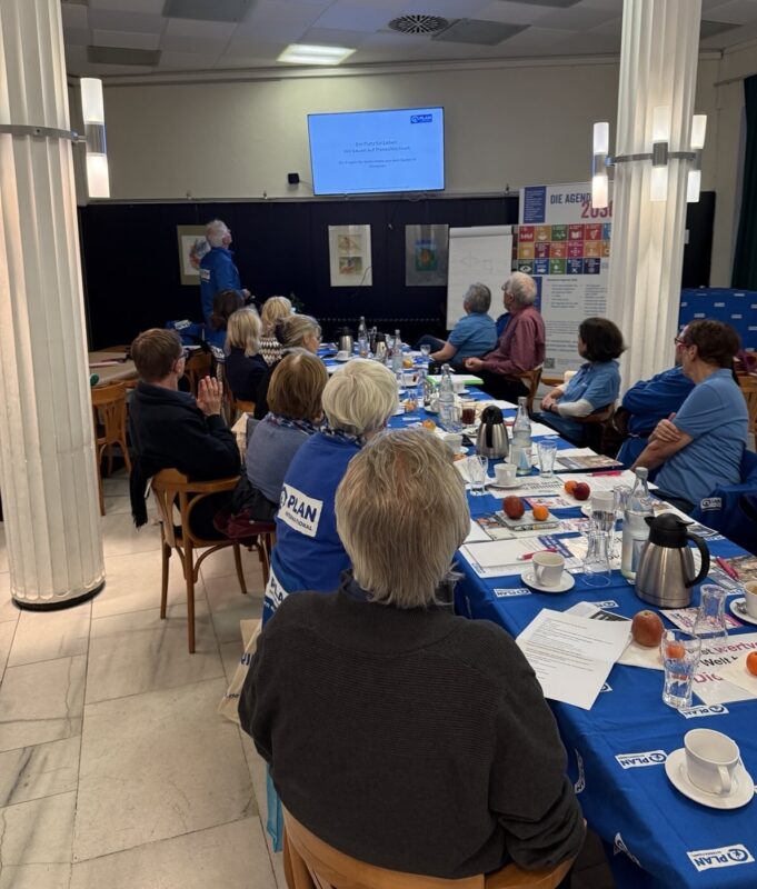Eine Gruppe von Menschen sitzt um einen langen Tisch mit blauen Tischtüchern, Papieren, Obst und Kaffeekannen. Eine Person steht vorne und zeigt eine Diashow auf einer Leinwand. Der Raum ist mit Säulen ausgestattet, und die Teilnehmer tragen blaue Hemden oder Jacken.