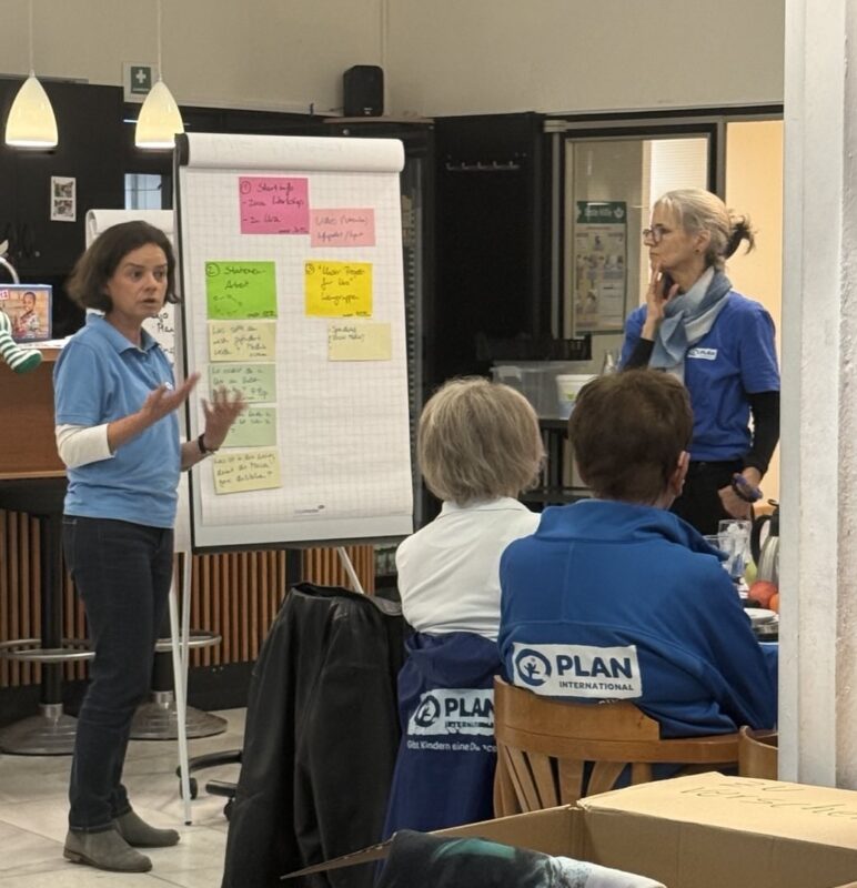 Zwei Frauen in blauen Hemden leiten einen Workshop. Die eine steht neben einem Flipchart mit bunten Notizen und gestikuliert, während sie spricht. Die andere steht da und hört nachdenklich zu. Mehrere Personen in passenden Hemden von Plan International sitzen an Tischen, die den Referenten zugewandt sind.