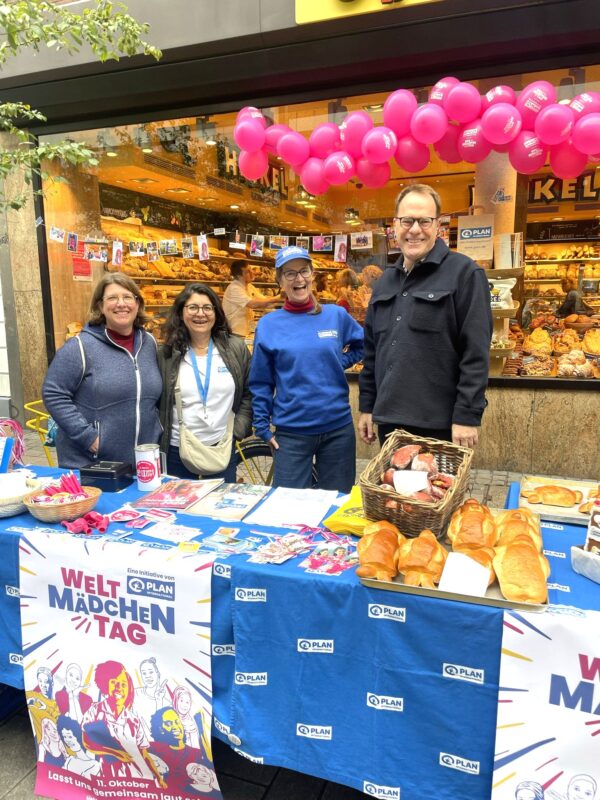 Vier Personen stehen lächelnd hinter einem bunten Stand mit Brot, Flyern und Plakaten, die für den Welt Mädchen Tag werben. Über ihnen hängen rosafarbene Luftballons, und im Hintergrund ist ein Bäckereiladen mit Gebäck zu sehen.