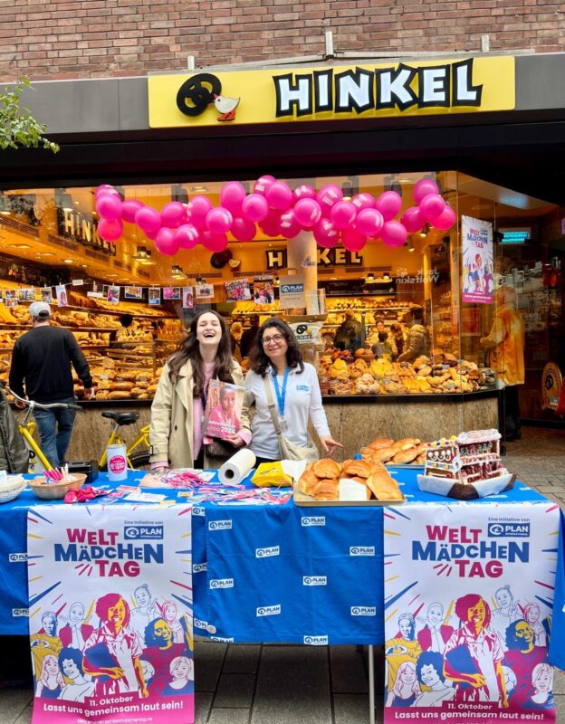Zwei Frauen lächeln hinter einem Tisch mit Brot und Snacks vor einer Hinkel-Bäckerei. Rosa Luftballons schmücken den Stand. Plakate mit der Aufschrift "Welt Mädchen Tag" werben für diesen Tag. Der Laden im Hintergrund ist voll mit Gebäck.