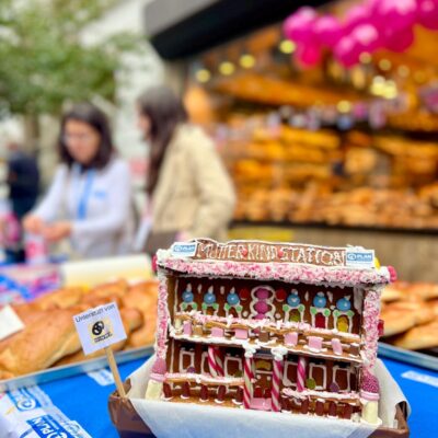 Ein Lebkuchenmodell mit der Aufschrift "Station Mutterkind" steht auf einem Tisch, der mit rosa, weißem und rotem Zuckerguss verziert ist. Dahinter stehen zwei Personen an einer Bäckereiauslage mit Brot und Gebäck, über denen rosa Luftballons schweben. Die Szene scheint bei einer Veranstaltung im Freien zu spielen.