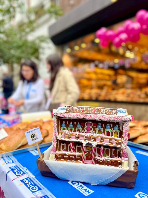 Ein Lebkuchenmodell mit der Aufschrift "Station Mutterkind" steht auf einem Tisch, der mit rosa, weißem und rotem Zuckerguss verziert ist. Dahinter stehen zwei Personen an einer Bäckereiauslage mit Brot und Gebäck, über denen rosa Luftballons schweben. Die Szene scheint bei einer Veranstaltung im Freien zu spielen.