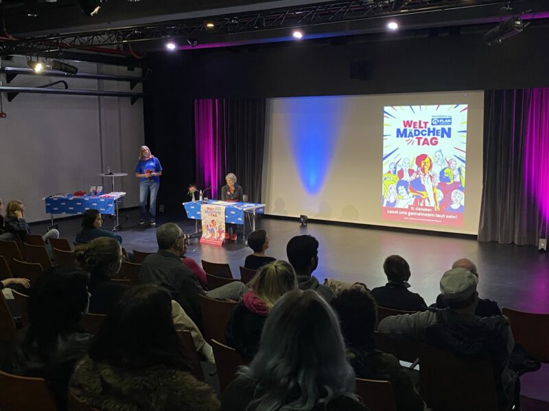 Ein kleines Publikum sitzt in einem dunklen Hörsaal vor einer Bühne, auf der eine Person steht und eine andere an einem Tisch sitzt. Auf einer Leinwand ist ein farbenfrohes Plakat mit dem Titel "Welt Mädchen Tag" zu sehen, auf dem illustrierte Mädchen und ein fetter Text abgebildet sind. Auf Tischen mit blauen Tüchern liegen Gegenstände.