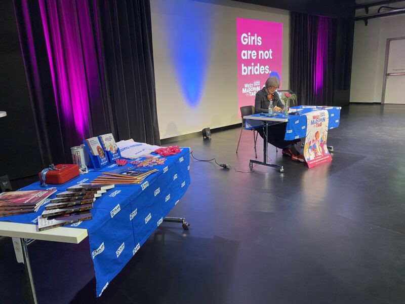 Eine Frau sitzt an einem Tisch auf der Bühne und liest aus einem Buch. Hinter ihr ist ein großer rosa Bildschirm mit weißer Schrift zu sehen: Mädchen sind keine Bräute. Auf den Tischen in der Nähe liegen Broschüren, Lesezeichen und Merchandising-Artikel aus. Die Atmosphäre ist hell mit lila und blauer Beleuchtung.