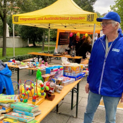 Ein älterer Mann in blauer Jacke und Mütze steht bei einer Veranstaltung im Freien an Tischen mit Spielzeug, Büchern und Spielen. Hinter ihm steht ein gelbes Zelt mit der Aufschrift Integrations- und Ausländerbeirat Dresden. Im Hintergrund sind andere Menschen und bunte Luftballons zu sehen.