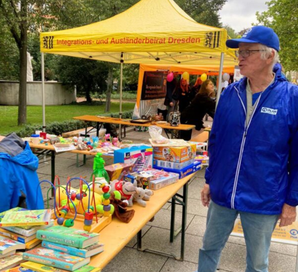 Ein älterer Mann in blauer Jacke und Mütze steht bei einer Veranstaltung im Freien an Tischen mit Spielzeug, Büchern und Spielen. Hinter ihm steht ein gelbes Zelt mit der Aufschrift Integrations- und Ausländerbeirat Dresden. Im Hintergrund sind andere Menschen und bunte Luftballons zu sehen.