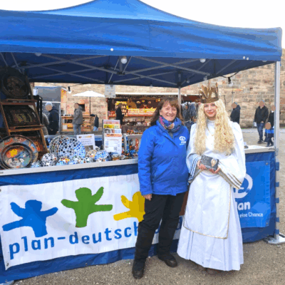 Zwei Frauen stehen vor einem blauen Stand. Die eine trägt eine blaue Jacke mit Plan-Logo, die andere ist als Engel oder Königin mit einem weißen Kleid, langer blonder Perücke und goldener Krone gekleidet. Buntes Kunsthandwerk und ein Transparent mit der Aufschrift "plan-deutschland" sind zu sehen.