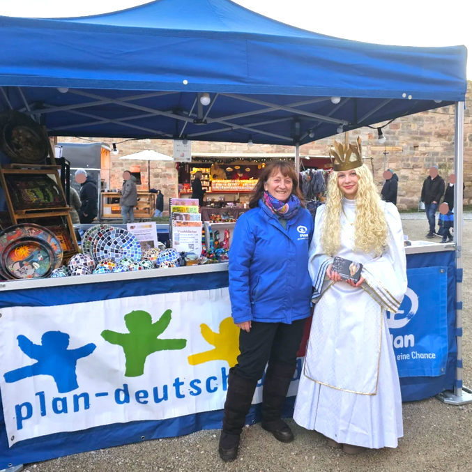 Zwei Frauen stehen vor einem blauen Stand. Die eine trägt eine blaue Jacke mit Plan-Logo, die andere ist als Engel oder Königin mit einem weißen Kleid, langer blonder Perücke und goldener Krone gekleidet. Buntes Kunsthandwerk und ein Transparent mit der Aufschrift "plan-deutschland" sind zu sehen.