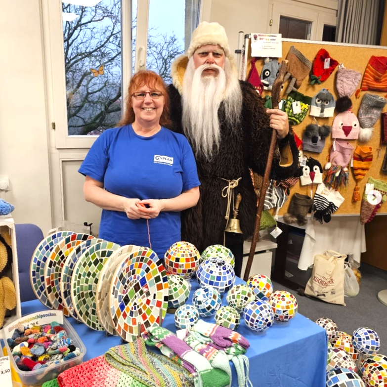 Eine lächelnde Frau in einem blauen Hemd steht an einem Kunsthandwerksstand mit bunten Mosaikkugeln und Strickwaren. Neben ihr steht ein Mann in einem langen braunen Gewand und weißem Bart, der einen Stab und eine Glocke hält. Hinter ihnen sind handgefertigte Hüte an ein Brett geheftet.
