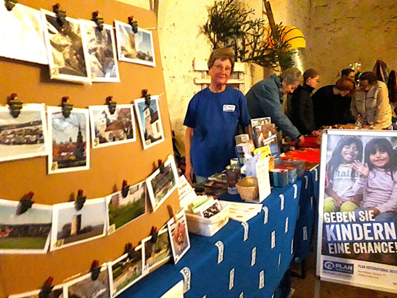 Eine Person in einem blauen Hemd steht hinter einem Tisch mit Broschüren und kleinen Gegenständen. Links sind Fotos an eine Tafel geklebt. Auf einem großen Schild mit zwei lächelnden Kindern steht Geben Sie Kindern eine Chance. In einem warm beleuchteten Raum stöbern die Leute an den Nebentischen.