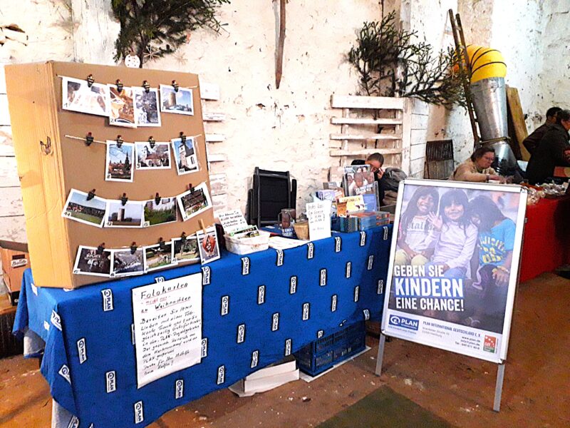 Ein Informationsstand mit einer blauen Tischdecke zeigt Fotos, Broschüren und Spendenmaterial. Ein großes Plakat zeigt drei lächelnde Kinder mit deutschem Text: Geben Sie Kindern eine Chance!", was so viel bedeutet wie: "Gebt Kindern eine Chance! Zwei Personen sitzen hinter dem Tisch.